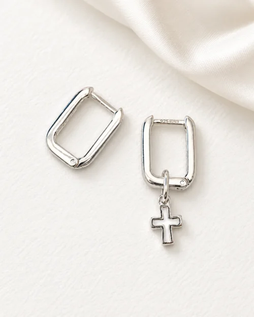 Pendientes Cosmos geométricos de aro rectangular en plata de ley 925, uno de ellos con colgante de cruz, sobre fondo color crema con tela satinada