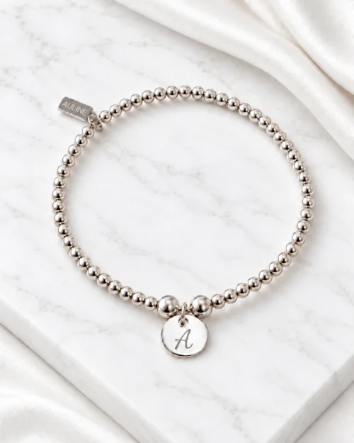 Pulsera Origen de bolas de plata de ley 925 con colgante redondo de 10 mm personalizable con inicial, sobre base de mármol color crema con tela satinada